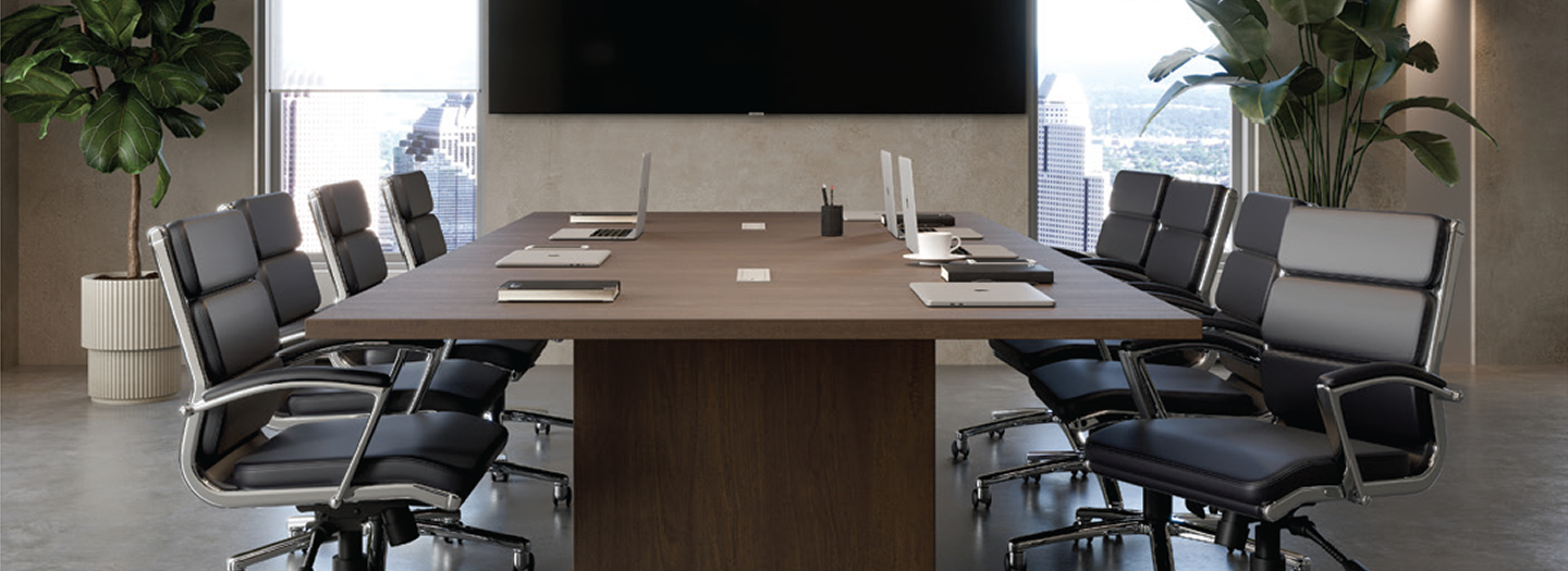 Medium wood laminate conference table with 8 black leather conference chairs featuring chrome bases and casters in a conference room with a large screen between floor to ceiling windows in a high rise building with large plants on either side.