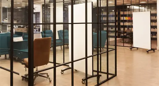 Freestanding mobile glass whiteboard on locking casters with sleek black frame positioned next to collaborative lounge seating in a bright modern open office. Perfect for flexible brainstorming, presentations, and agile team meetings.