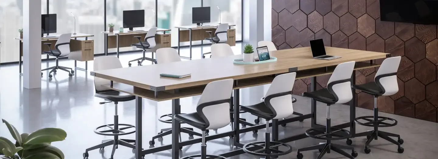 Industrial manufacturing collaborative workspace with long height-adjustable wood table, white swivel counter stools, stand alone desks with monitors and storage for focused work, hexagonal wood wall, and polished concrete floors for team huddles and project reviews.