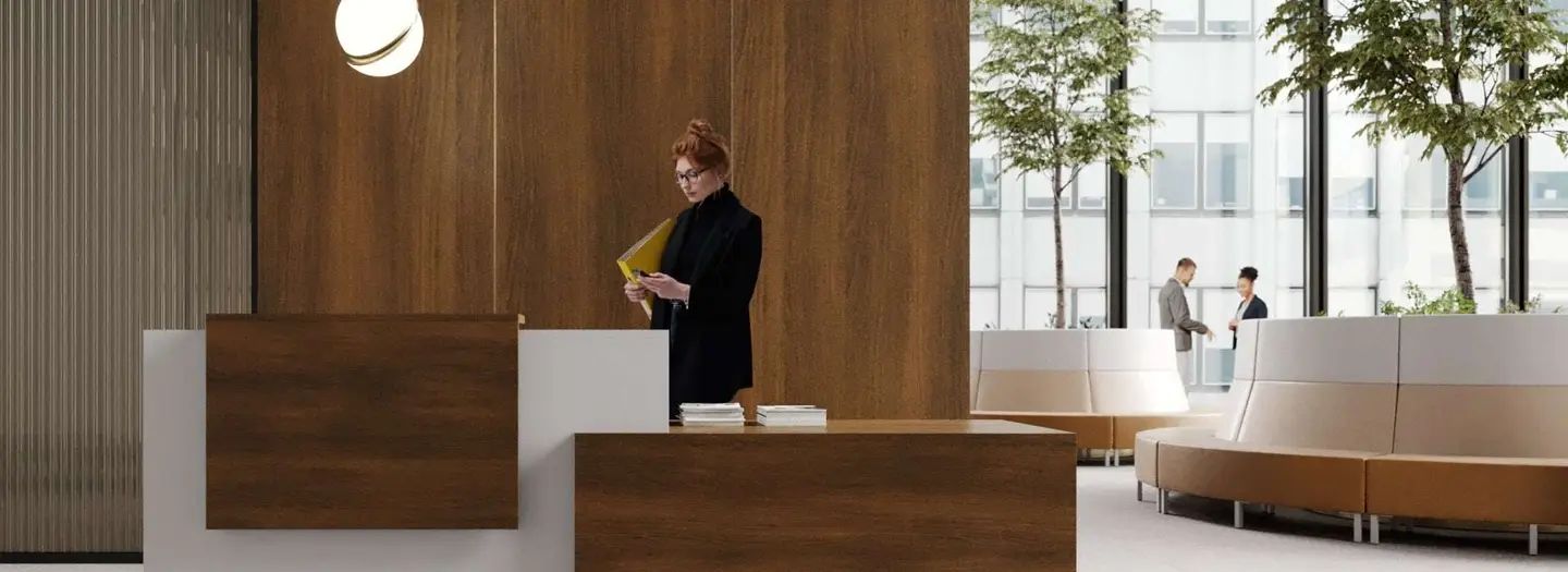 Hero: Welcoming corporate reception with white-and-wood laminate desk, employee checking her phone, warm wood accent wall, and two round lounge seating groups with large trees for visitor waiting and donor meetings.