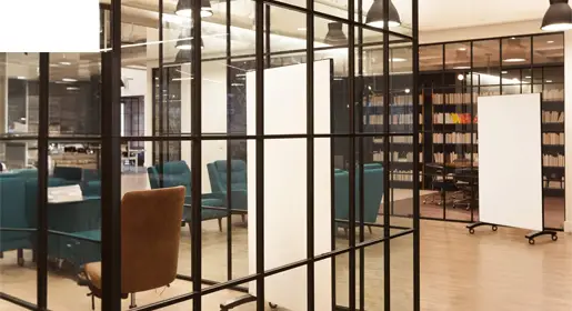 Freestanding mobile glass whiteboard on locking casters with sleek black frame positioned next to collaborative lounge seating in a bright modern open office. Perfect for flexible brainstorming, presentations, and agile team meetings.