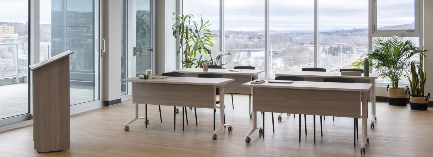 Bright, naturally lit training space with four light wood folding tables on wheels, black ergonomic chairs, podium, and large potted plants overlooking urban landscape. Ideal for reconfigurable classrooms and all-day seminars.