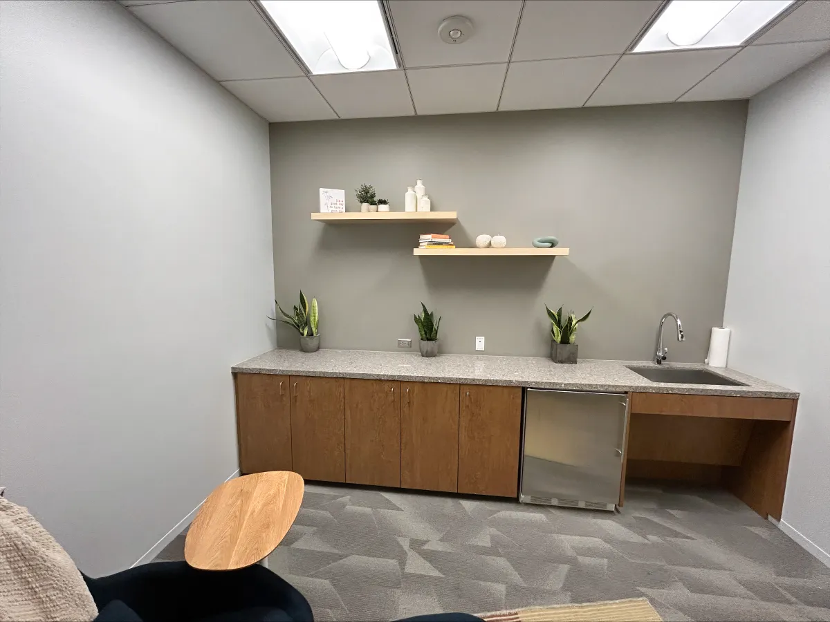 Modern breakroom storage cabinet from OFC CONNECT Series in warm oak wood laminate with granite-look laminate countertop, undermount sink, mini fridge space, floating shelves, and live plants – complete corporate kitchenette solutions by Office Furniture Connection Houston TX.