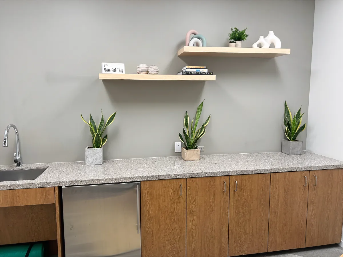 Stylish coffee station credenza from OFC CONNECT Series in natural oak wood laminate featuring granite-look countertop, floating display shelves, under-counter refrigerator, and decorative plants – upscale office breakroom storage and beverage center by Office Furniture Connection Houston TX.