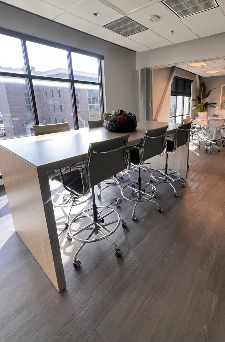 Tall collaborative standing-height cafe table from OFC CONNECT Series in light oak wood laminate with chrome adjustable stools, and natural window light – modern open office break area and huddle space furniture by Office Furniture Connection Houston TX.