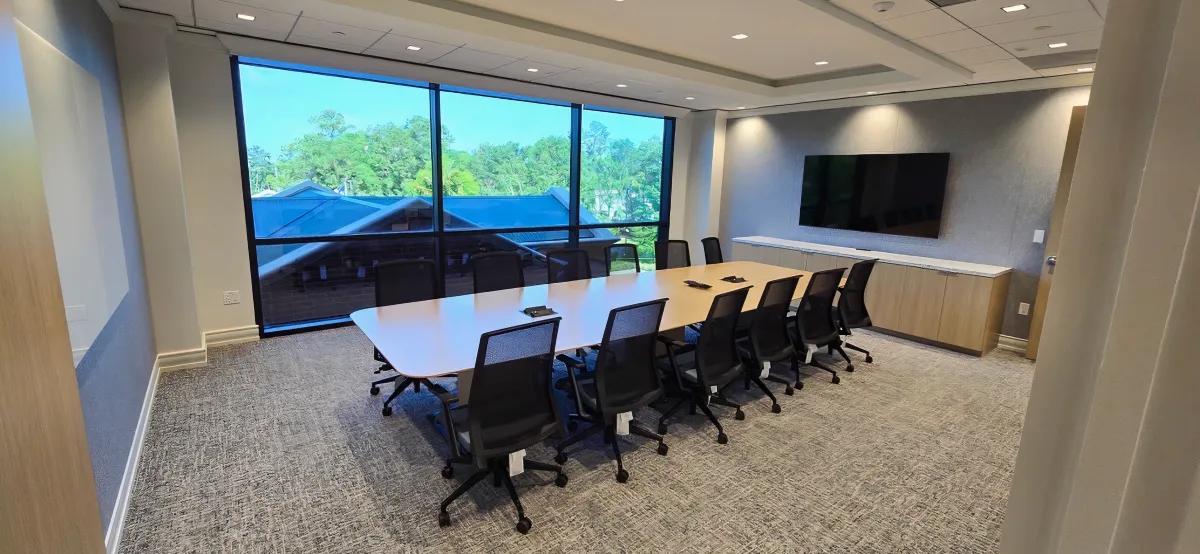 12-foot boat-shaped conference table from OFC CONNECT Series in light oak wood laminate with integrated power grommets, surrounded by 12 black ergonomic mesh conference chairs, matching credenza, and wall-mounted TV – premium modern boardroom solutions by Office Furniture Connection Houston TX.