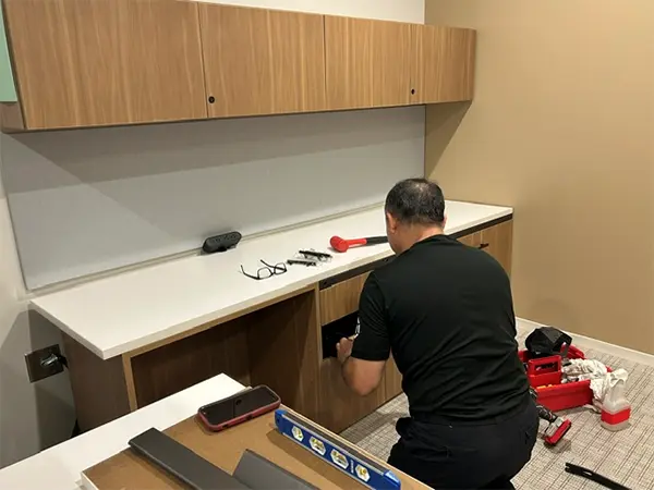 Professional furniture installer assembling OFC CONNECT custom millwork wood-grain office cabinetry with overhead storage and white countertop for precise commercial delivery and installation service.