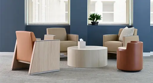 Modern healthcare waiting room with beige and tan upholstered lounge chairs around a round wood laminate table, tissue box, and relaxing blue wall with large windows for a calming patient experience.