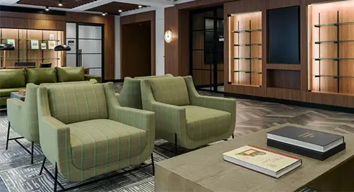 Elegant lobby seating area with large green plaid overstuffed chairs, green leather sofa, wood coffee tables, and illuminated built-in shelving for upscale hotel guest relaxation and meetings.