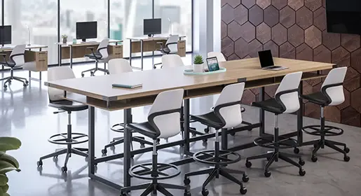 Industrial manufacturing collaborative workspace with long height-adjustable wood table, white swivel counter stools, stand alone desks with monitors and storage for focused work, hexagonal wood wall, and polished concrete floors for team huddles and project reviews.