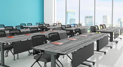 Large, high-ceiling training room with 16 gray wood-grain flip-top tables with modesty panels and casters, arranged in rows, black mesh chairs, acoustic ceiling baffles, and turquoise accent wall. Ideal for scalable corporate training and certification programs.