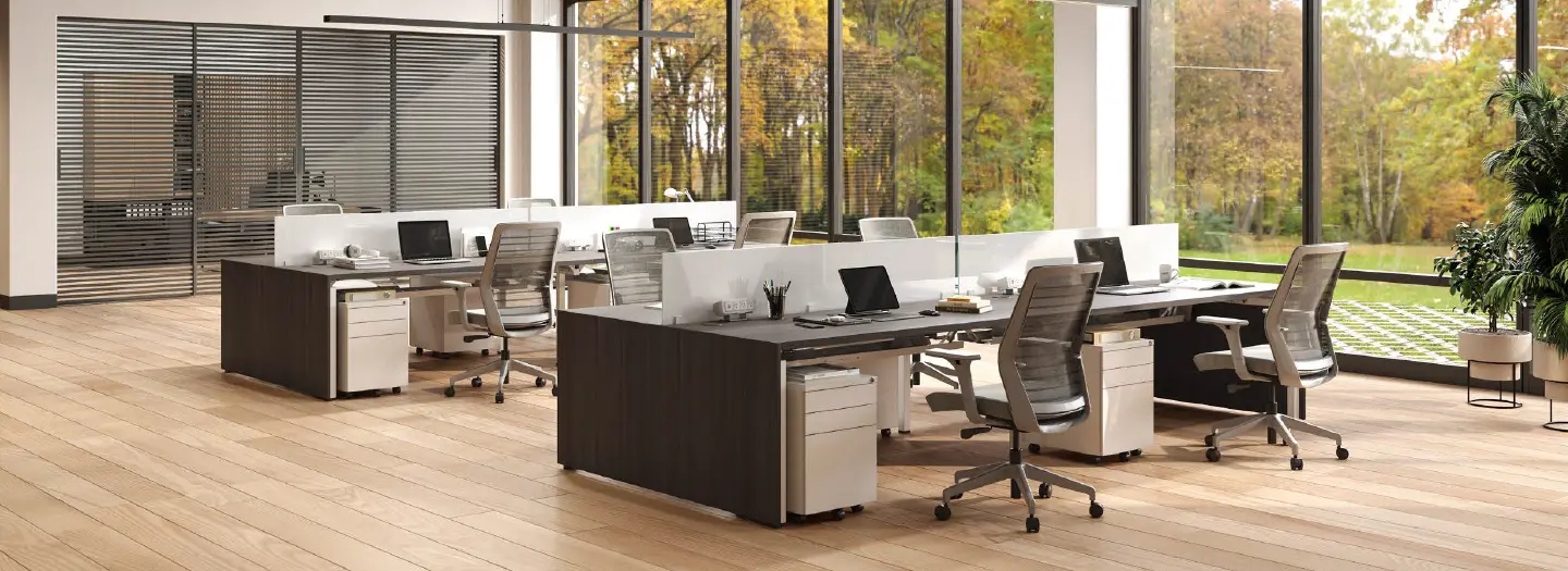 A bright open office space with floor to ceiling windows looking out onto a wooded area during the fall. Two groups of benching stations with 4 pods each sit in the middle of the space. Storage includes a box/box/file and low barrier on the worksurface is between each station.