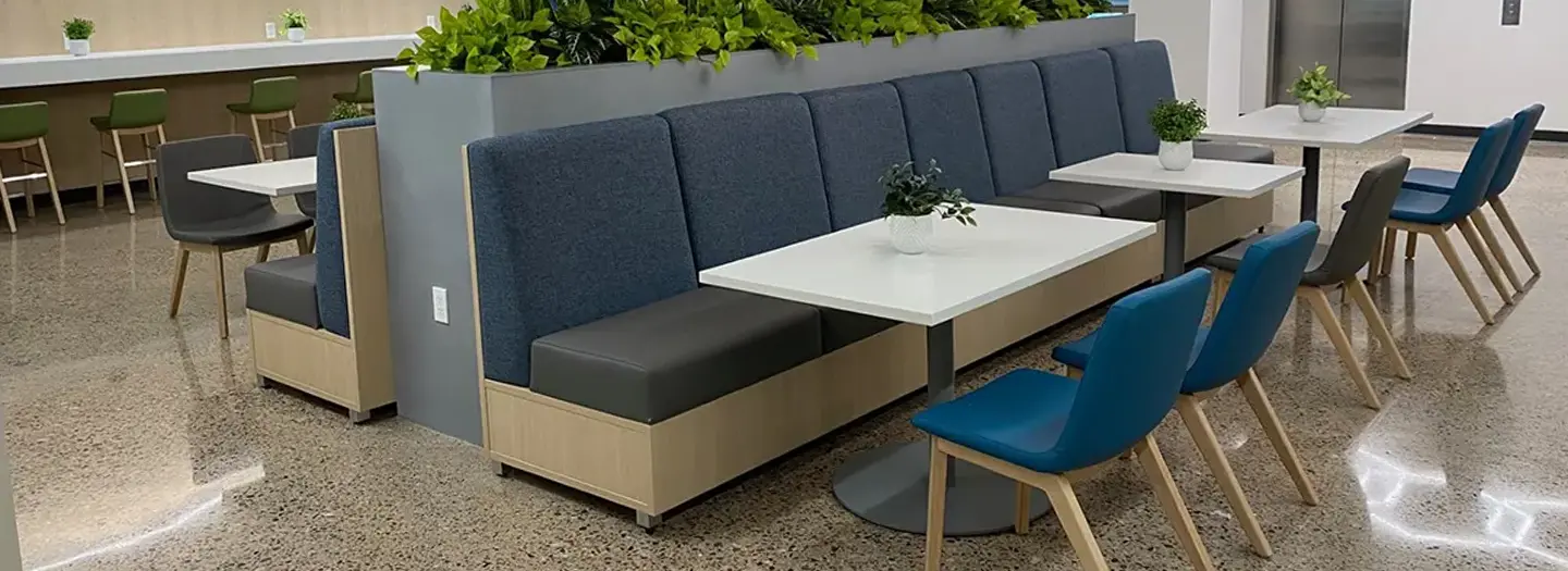 A Office Furniture Connection project with three new white laminate cafe tables that each seat four, lined up along a length of benches on one side and new blue cafe chairs along the other side. A tall planter sits behind the benches and the same layout of breakroom furniture is matched on the other side of the planter.