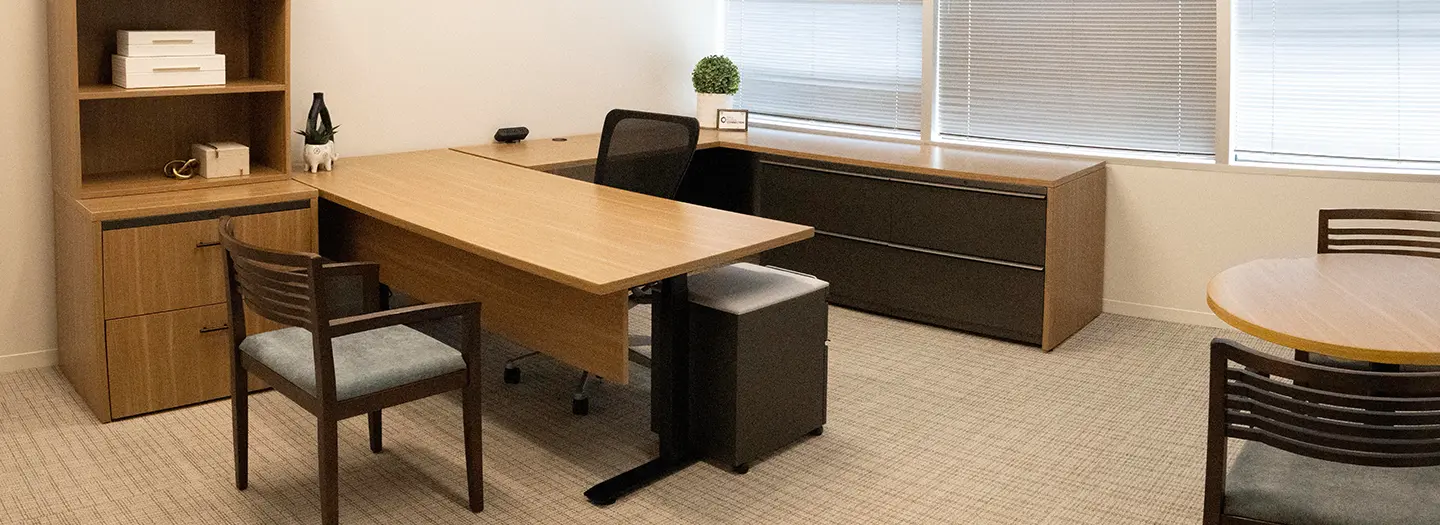 Neutral colored private office with u-shaped sit stand desk in honey wood laminate with (2) 2 drawer black lateral files and a box/file mobile ped. A black task chair with three guest chairs and 36" round laminate table. A matching two drawer lateral file with stacked bookcase.