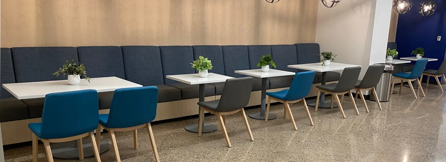 A Office Furniture Connection project with six new white laminate cafe tables with black ciruclar bases that each seat two or four, lined up along a length of benches on one side and new blue cafe chairs along the other side. Each table has a small plant.