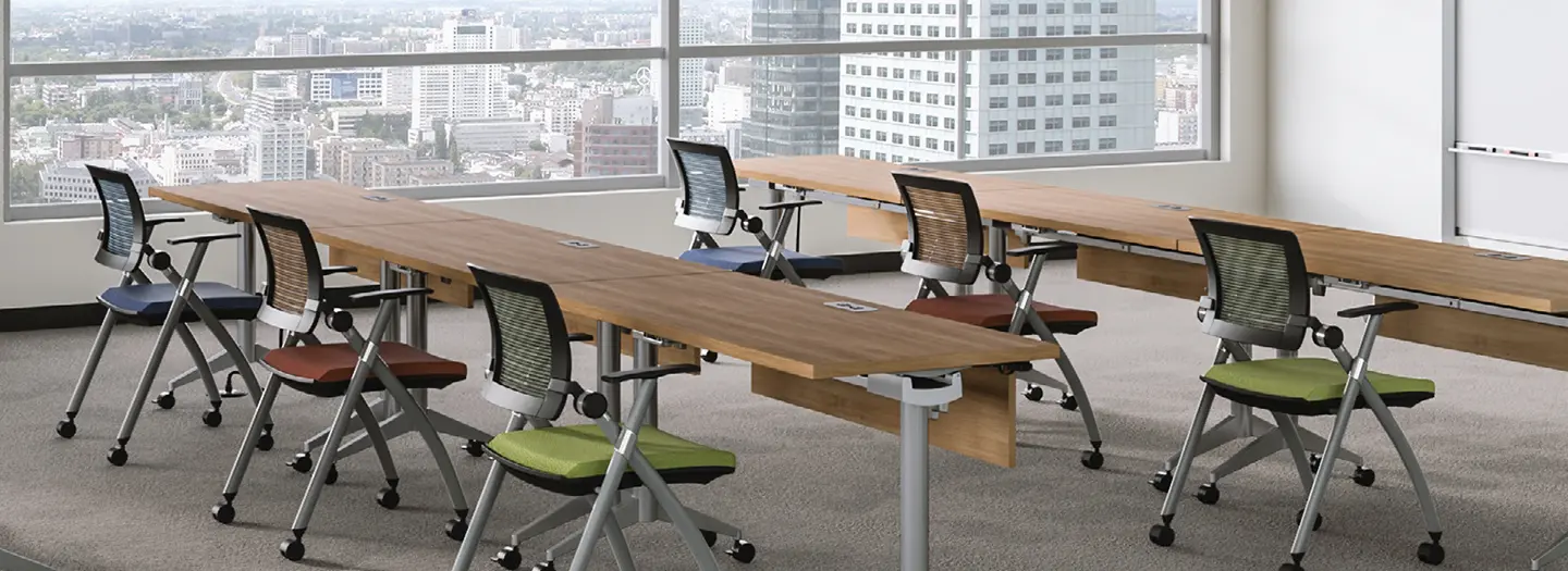 training room in a skyscraper wtih floor to ceiing windows includes 6 flip top training tables in wood laminate with modesty panels and metal bases. Training chairs are colorful blue, red, and citrus green fabrics with mesh backs and wheels. All are facing a marker board at the front.