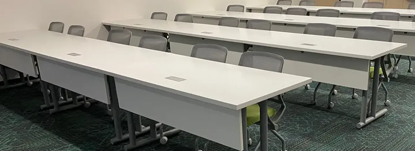 Four rows of flip top training tables with modesty panels and laminate tops are made even more useful with the addition of data and power ports. Two light-weight training chairs with wheels pull up to each table.