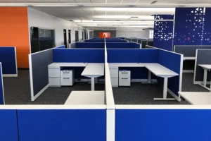 Close-up of remanufactured Knoll Morrison cubicles with 42H vibrant blue panels, electric height-adjustable desks, mobile pedestals, and powered spines in a carpeted office with orange walls. Affordable office redesign solution.
