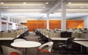 Open office featuring remanufactured Knoll Morrison cubicles with 42H beige panels, ergonomic chairs, white worksurfaces, and orange glass partitions around a breakout table. Great for modern hybrid workspaces.