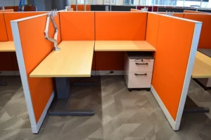 Remanufactured Knoll Morrison workstation featuring bright orange 48H fabric panels, L-shaped wood-grain adjustable height desk, dual monitor arm, and 6/12 mobile pedestal in a carpeted open office. Ideal for bold, cost-effective office layouts.