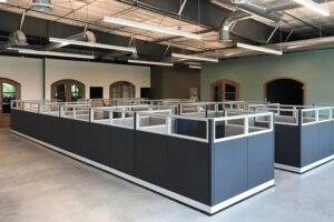 Large group of Knoll Morrison remanufactured6x6 workstations with 56H charcoal panels and glass stackers arranged in pods inside a high-ceiling warehouse with exposed ducts. Turnkey solution for 50+ employee office layouts.
