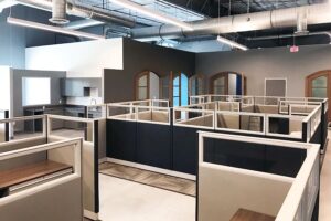Remanufactured Knoll Morrison office cubicles with 56H navy and beige panels, framed glass accents, wood laminate worksurfaces arranged in a cluster within an industrial warehouse featuring arched doors and exposed HVAC ducts. Ideal for budget-friendly, collaborative workspaces.