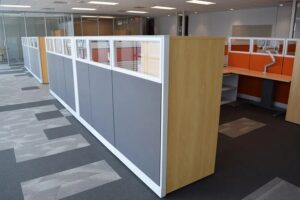 Knoll Morrison remanufactured office cubicles with 56H gray and orange fabric panels, glass stackers, light wood wardrobe storage towers, and height-adjustable desks with monitor arms in a bright office space. Perfect for large-scale, budget-friendly office installations.