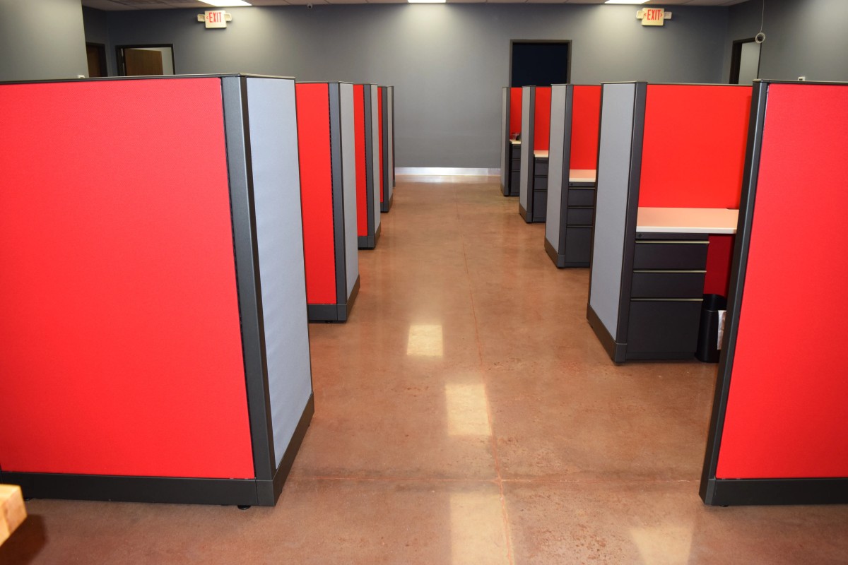 Two rows of remanufactured cubicles with bold red and blue 56H fabric panels, white worksurfaces, and box/box/file pedestals arranged in an open plan with polished concrete floors. Affordable, high-quality workstations ready for immediate delivery and installation.