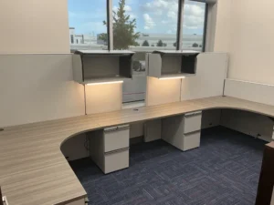 Two Knoll Morrison remanufactured shared cubicles with light gray 64" panels for overhead storage and tasklight and 48" low panels, medium-tone wood laminate corner worksurfaces with grommets and file/file pedestals