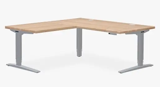 OFD corner L-shaped height-adjustable sit-stand desk in natural oak laminate with silver 3-stage legs, integrated power grommets, and smooth electric lift – ergonomic standing corner workstation solution for executive and home offices by Office Furniture Connection Houston TX.