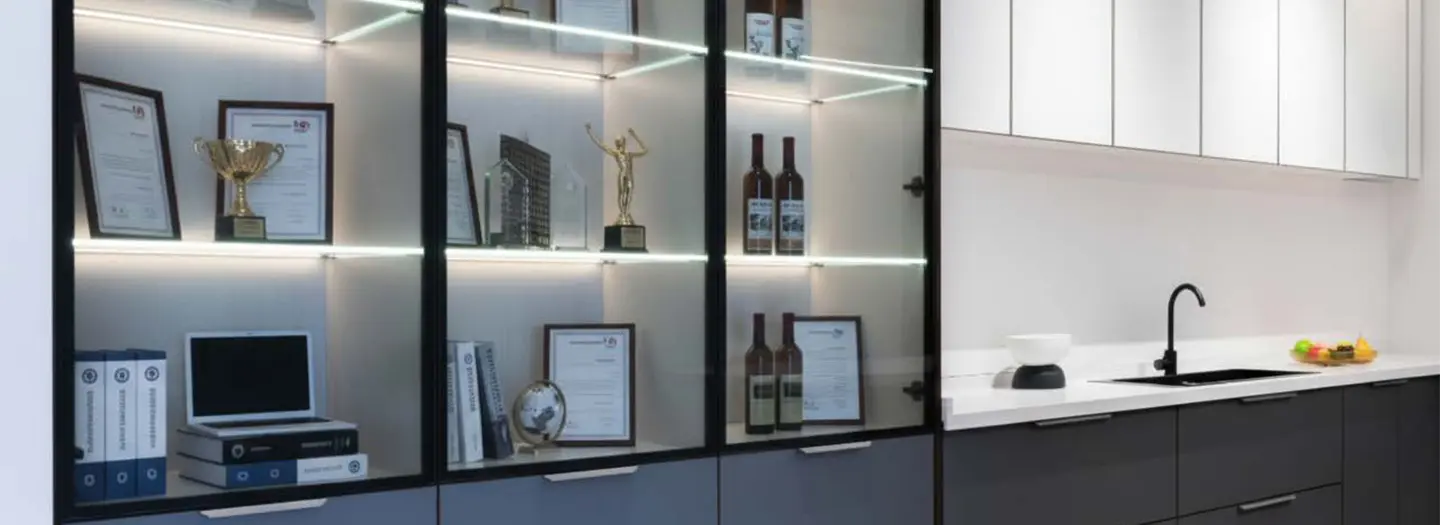 Elegant black-framed glass display cabinet with integrated LED shelf strip lighting showcasing trophies, awards, and wine bottles – luxury executive office accent illumination by Office Furniture Connection Houston TX.