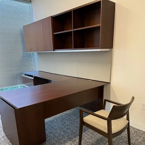 An executive office with used office furniture or casegoods including medium wood tone L-shaped desk with tackboard in beige and open and closed storage above. Filing below and wood upholstered guest chair.