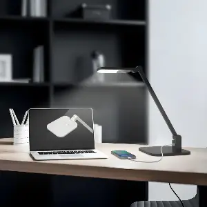 MOD Nebula Desk Lamp in Black with slanted arm highlighting a worksurface with other desk accessories and a black bookcase in the background.